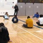 A group of students sit on a school gym floor while a teacher stands in front of a white board giving a lesson.