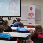 A group of students sit in a conference room with a student panel at the front of the room. An adults provides group facilitation.